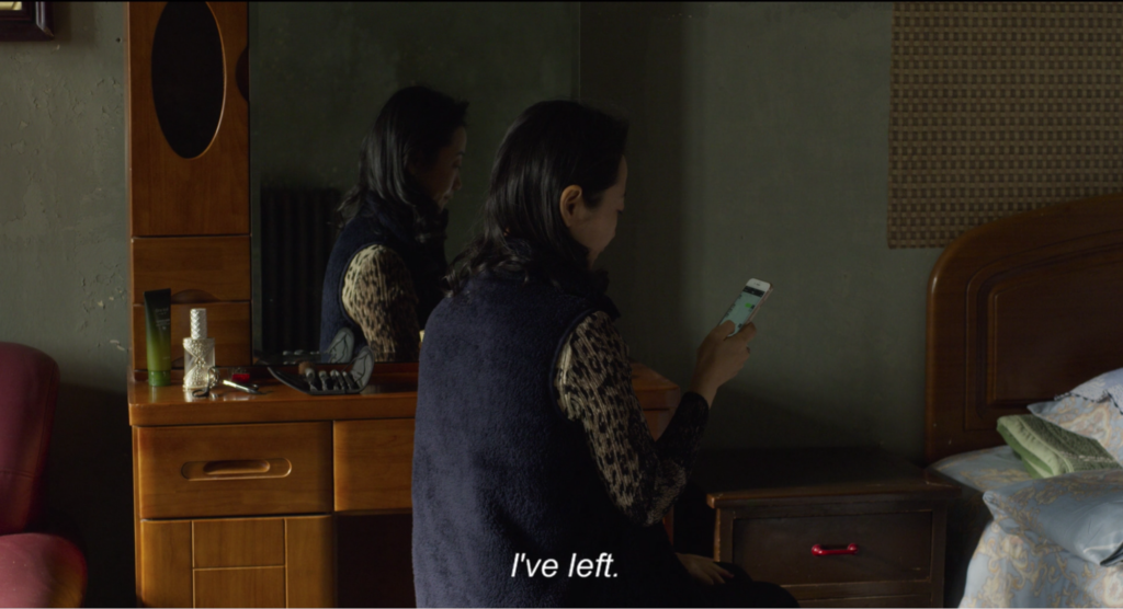 Resembles a still from a film. A woman with black, shoulder-length hair dressed in a navy-blue vest and a leopard-print long sleeve stands in a bedroom. She looks at her iPhone in her right hand. she faces away from the camera. Her left side is reflected by the vanity mirror in the background. The subtitle positioned at the bottom center of the still says "I've left." in white san-serif text, black border italics. 