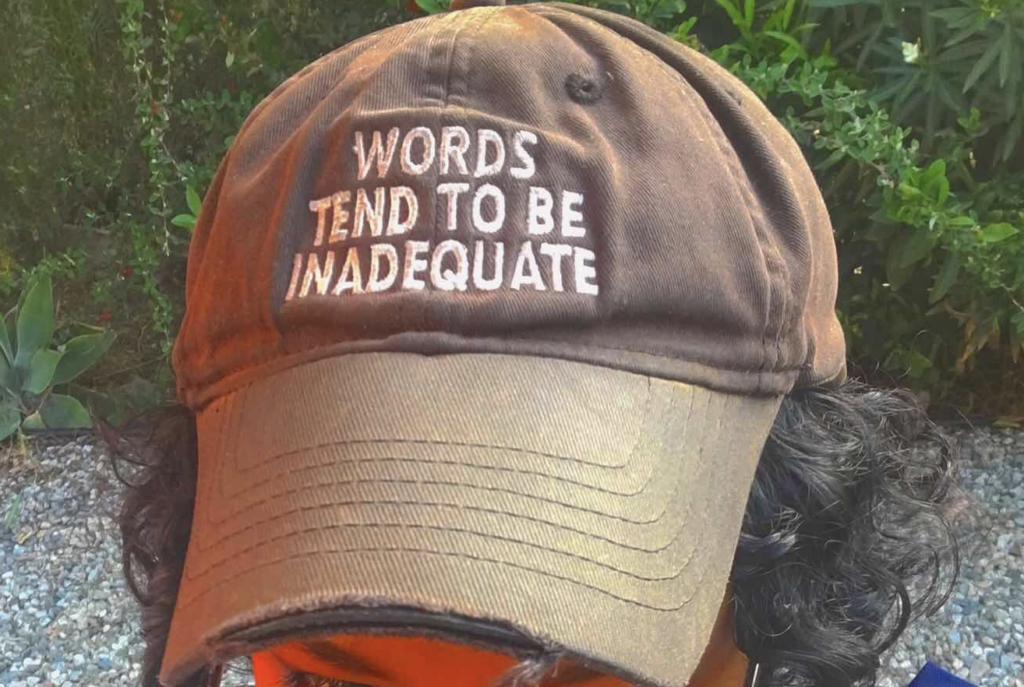 A close-up view of a person wearing a brown baseball cap that is embroidered in white san-serif text with the words "WORDS TEND TO BE INADEQUATE." The brim of the cap is slightly torn. The person sporting the cap is unidentifiable except for the black curly hair that overhangs from the sides of the cap. 