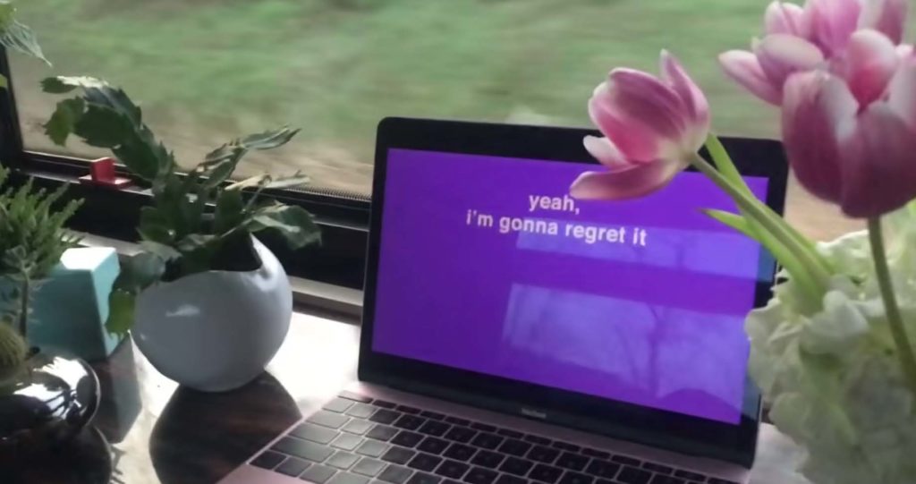 A laptop with a purple screen and text that reads "yeah, i'm gonna regret it" sitting in front of a window looking on a grassy field.